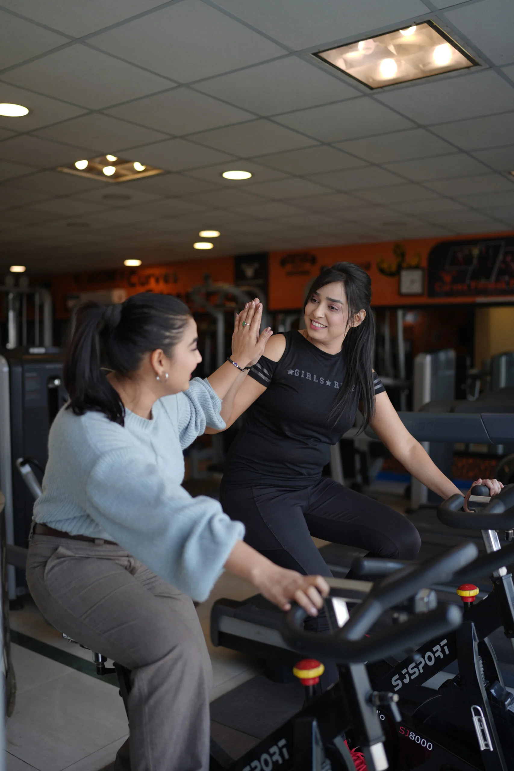 Two women smiling and high-fiving while exercising on stationary bikes in a brightly lit gym.