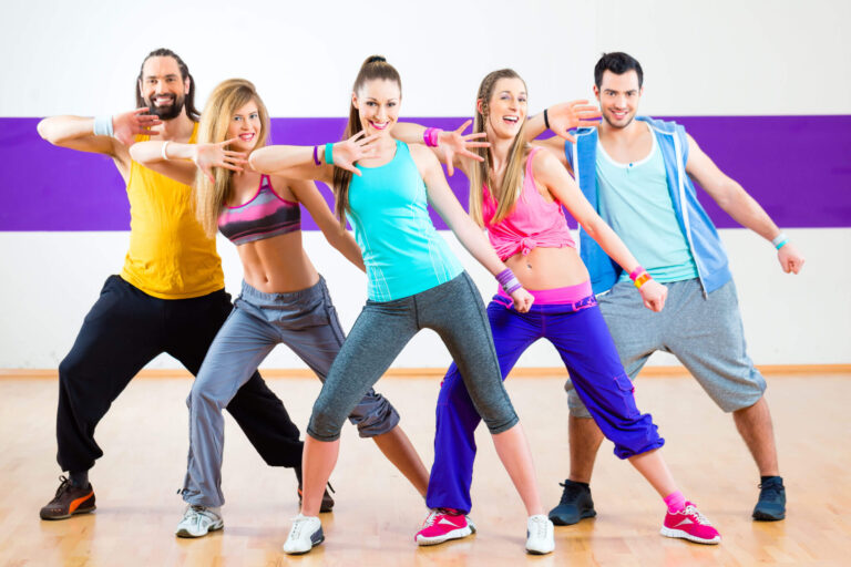 Group of energetic dancers in colorful outfits doing Zumba in a bright studio with wooden floors.